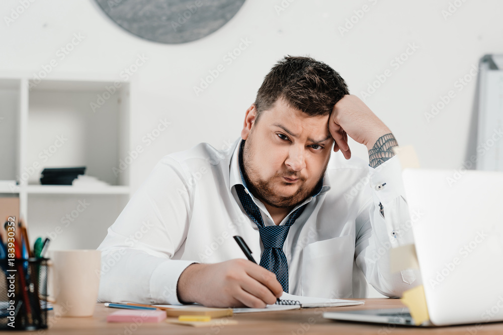 upset overweight businessman working with notepad and laptop in office upset overweight businessman working with notepad and laptop in office
