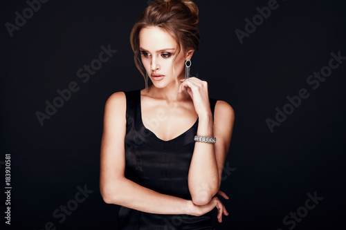 Attractive amazing sexy blonde woman with glamorous makeup, wearing silky black dress and chic jewelry, posing in studio with dark background