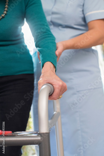 Senior Woman's Hands On Walking Frame With Care Worker In Background