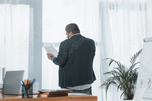 Bild auf Leinwand back view of overweight businessman in suit reading document at window in office
