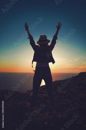 Carefree womans silhouette hands up in sunset on cliff
