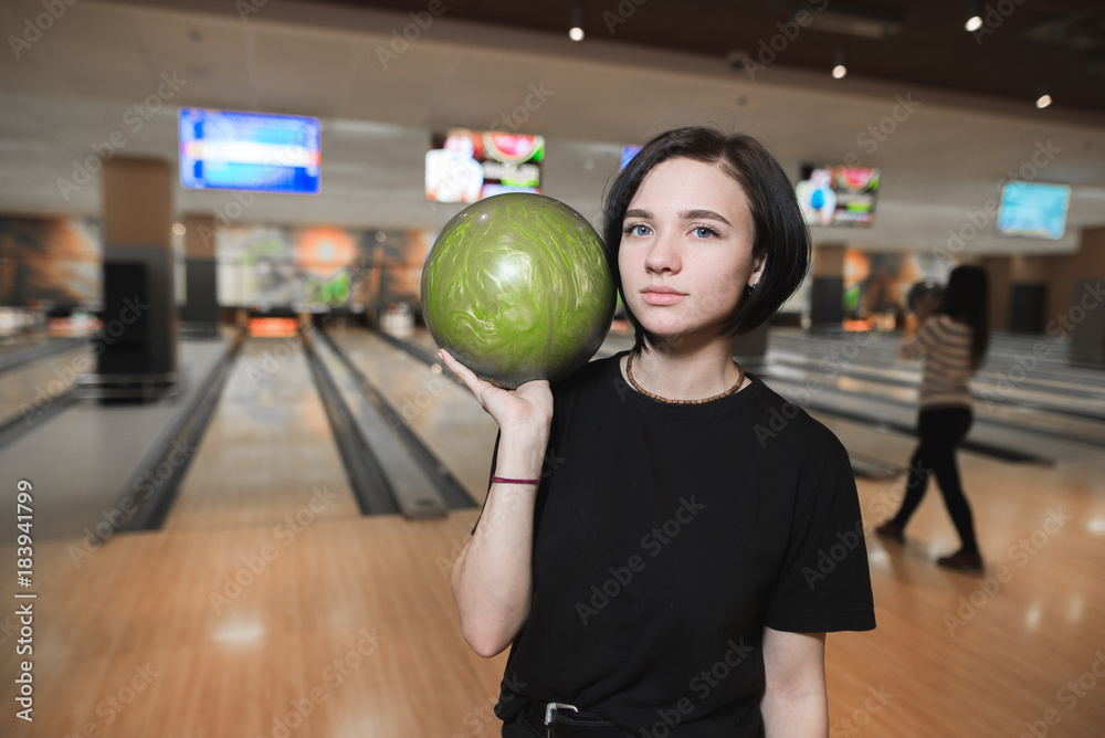 Beautiful young girl with green ball for bowling. The girl stands against the background of the bowling lanes and looks at the camera Beautiful young girl with green ball for bowling. The girl stands against the background of the bowling lanes and looks at the camera