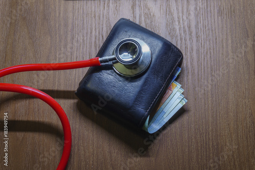 Conceptual image of diagnosing personal financial strength. Stethoscope on wallet with partly visible currency notes over brown surface. Selective focus; dimmed study lamp.