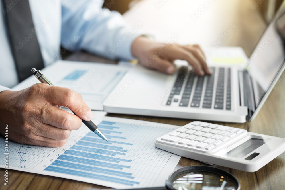 Elegant businessman working on his laptop in a coffee shop, analyzing data and graph, using calculator calculate, young man typing on computer sitting at wooden table Elegant businessman working on his laptop in a coffee shop, analyzing data and graph, using calculator calculate, young man typing on computer sitting at wooden table