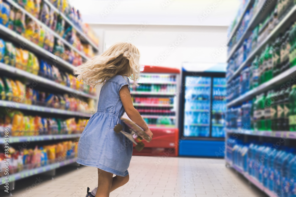 little kid with skateboard standing in supermarket with shelves behind