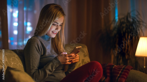 Beautiful Young Woman Sitting on Her Sofa in a Living Room. She Uses Smartphone with Interest. It's Evening, Big City is Seen in the Window.