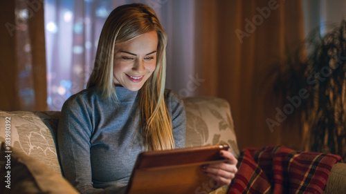 Beautiful Young Woman in Her Living Room. She is Sittin on a Sofa and Uses Tablet Computer. Behind Her Big City is Seen in the Window. Warm amdient.