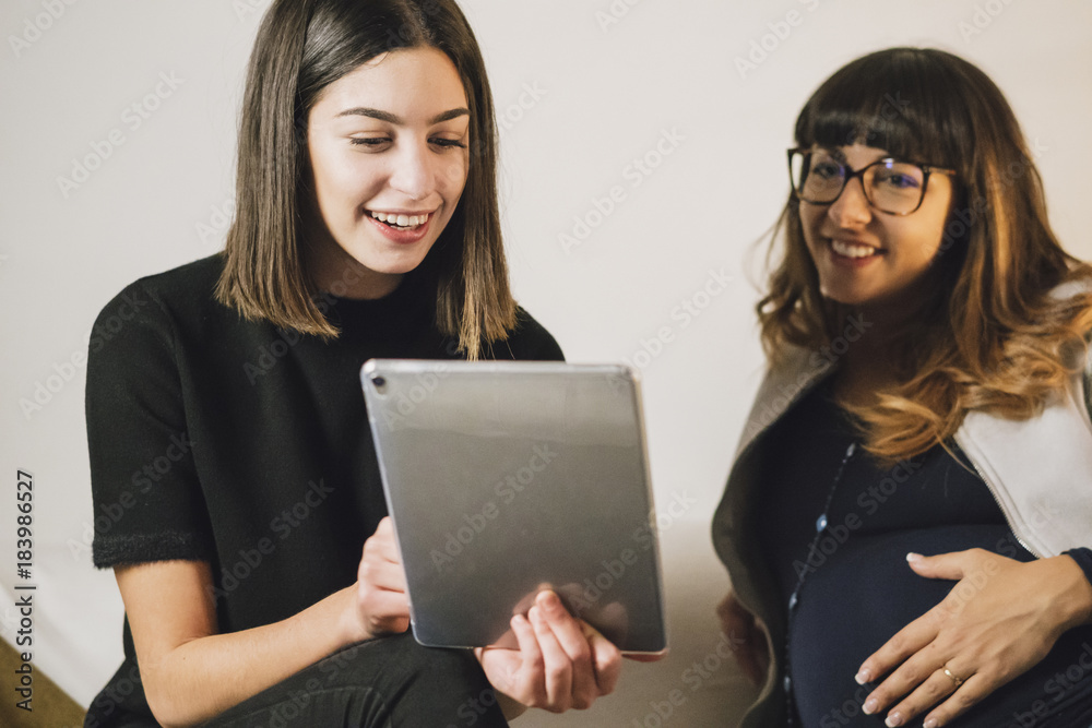 Pregnant woman and her friend look at a tablet on a couch Pregnant woman and her friend look at a tablet on a couch