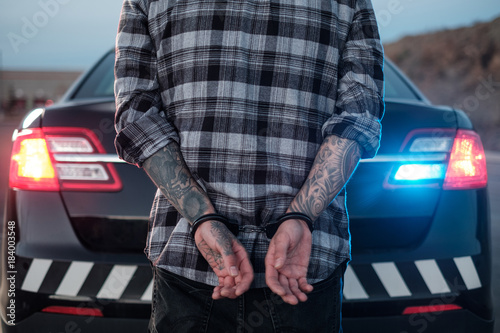 Tattooed arms & hands of a suspect in handcuffs behind a police cruiser