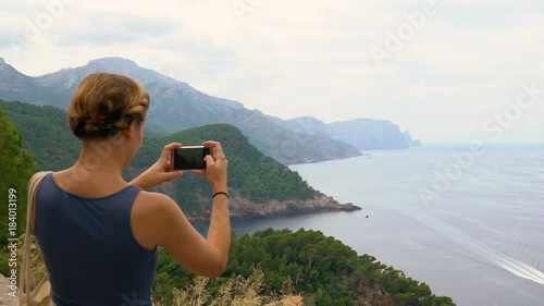 Young happy woman takes smartphone pictures on top of the mountain clifff from the beautiful ocean landscape photo