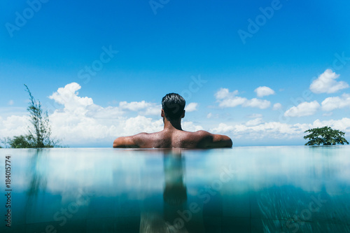 Topless man at infinity pool's edge against sky with fluffy clouds in a hotel