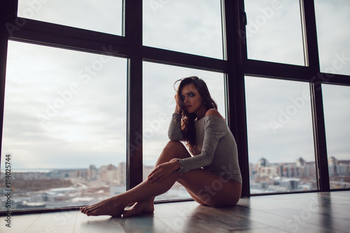 Sexy woman in gray bodysuit sits on the floor near the panoramic window.