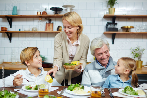 Grandmother giving boiled corn to her grandson by breakfast with mature man and little girl near by