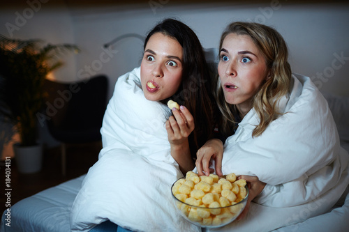 Curious girls eating corn rolls from bowl while watching captivating movie at night