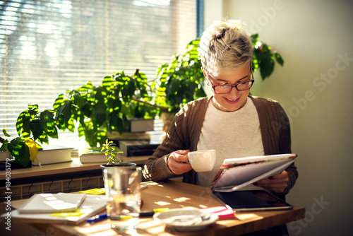 Charming cheerful satisfied middle age short hair woman sitting at the table in the morning at home and drinking a coffee while reading students tests or business statistics.