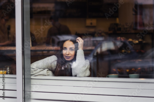 portrait of young beautiful cute brunette girl in grey sweater looking at the camera in coffee shop throuth glass window
