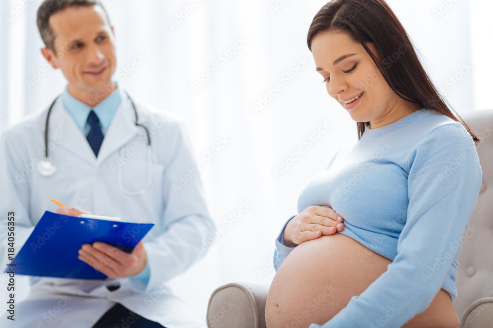 Feeling happiness. Beautiful future mommy bowing her head while watching her belly and sitting in semi position Feeling happiness. Beautiful future mommy bowing her head while watching her belly and sitting in semi position