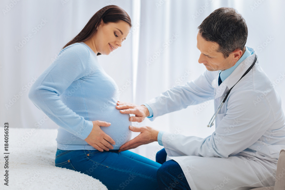 Fototapeta premium Let me see. Competent medical worker examining baby bump and bowing head while sitting opposite his patient
