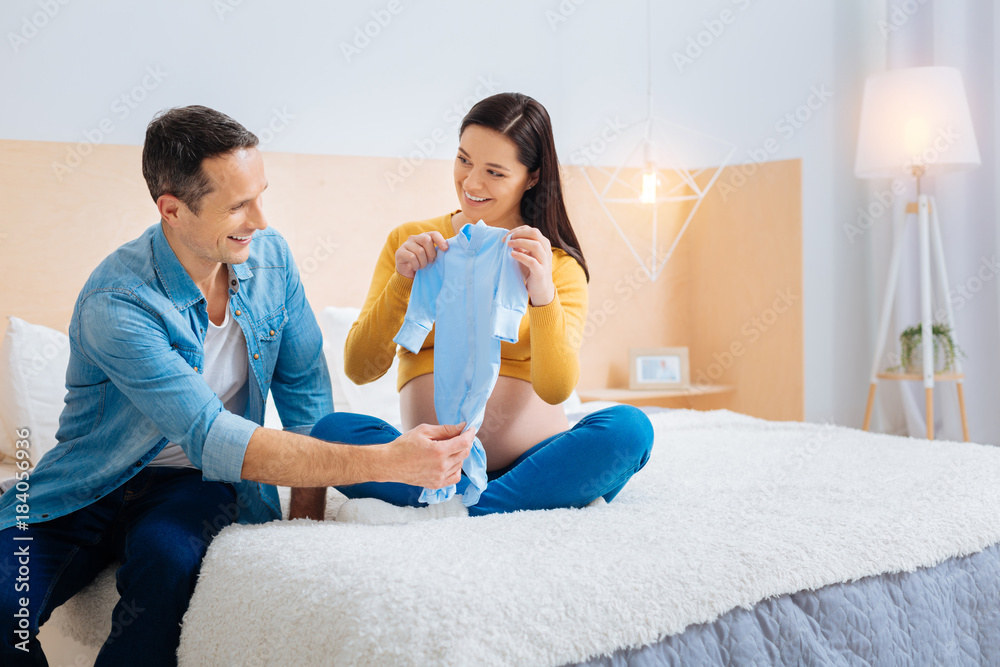 Good quality. Delighted man keeping smile on his face and holding romper while talking to his wife Good quality. Delighted man keeping smile on his face and holding romper while talking to his wife