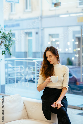 Free Download Vertical portrait of charming stylish female client with smartphone hands drinking cup of cappuccino and waiting for make up artist at modern interior beauty parlor or cafe. - GFXLoad