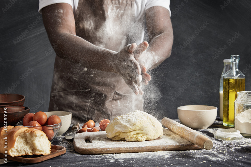 Φωτογραφία Professional male cook with dark skin, wears apron, sprinkles dough with flour, preapares or bakes bread at kitchen table, has dirty uniform, isolated over black chalk background