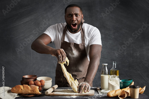 Shocked dark skinned professional male baker prepares dough at kitchen, kneads dough, can`t understand why it is sticky, needs more flour to make thick, isolated over black chalk background.