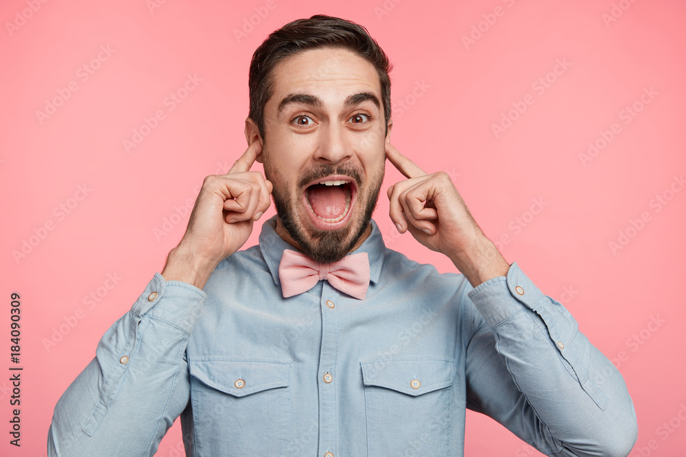 Portrait of bearded young male has cheerful expression, plugs ears, makes fun, isolated over pink background. Man with pleasant appearance hears loud music on disco party. I don`t hear anything Portrait of bearded young male has cheerful expression, plugs ears, makes fun, isolated over pink background. Man with pleasant appearance hears loud music on disco party. I don`t hear anything
