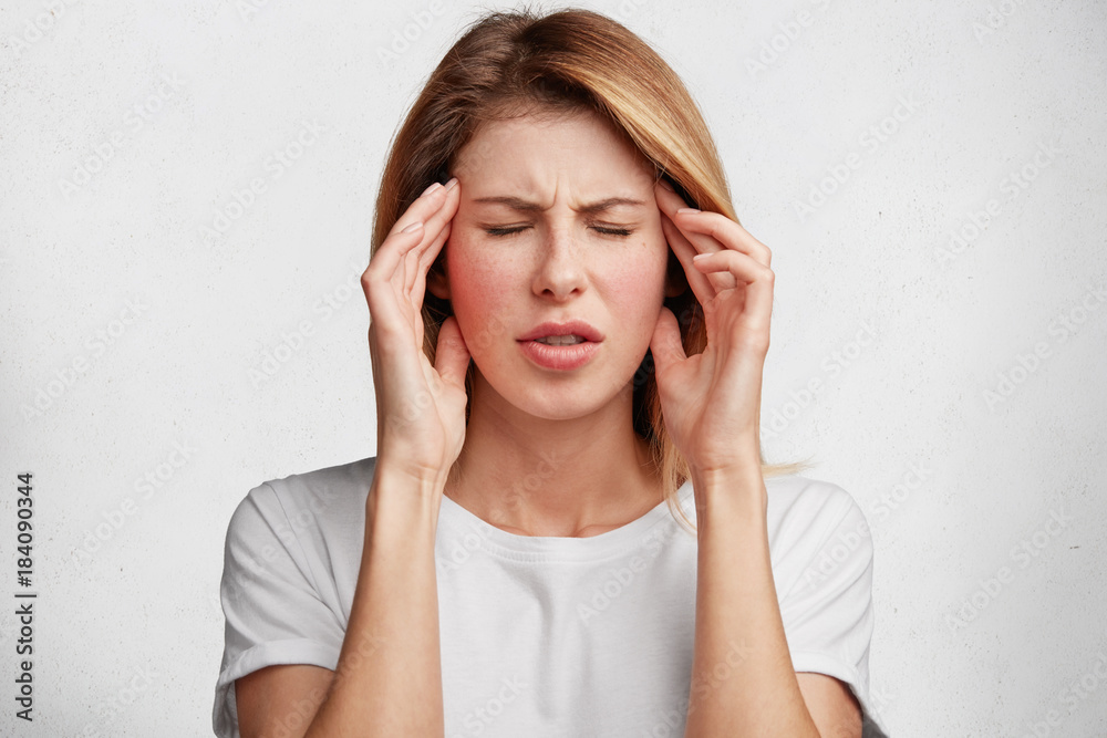 Portrait of fatigue middle aged female has terrible headache, keeps hand on temples, feels pain and frustration, closes eyes, has unhappy expression, isolated over white concrete background.