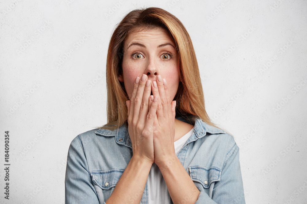 Photo of shocked middle aged beautiful woman covers mouth with hands, dressed in fashionable denim shirt, tries to control emotions, being afraid of something, isolated over white background Photo of shocked middle aged beautiful woman covers mouth with hands, dressed in fashionable denim shirt, tries to control emotions, being afraid of something, isolated over white background