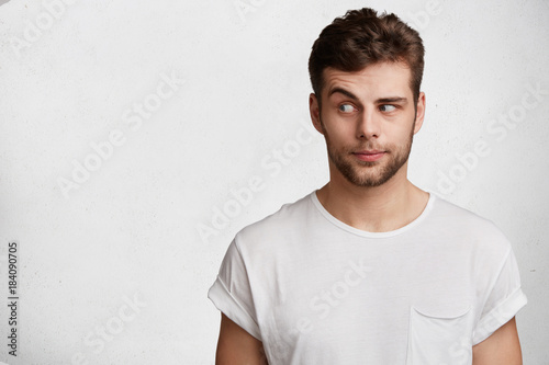 Young bearded attractive male with skeptic look, raises eyebrow in bewilderment, has some doubts and uncertainty, looks curiously aside, isolated over white background with copy space for text