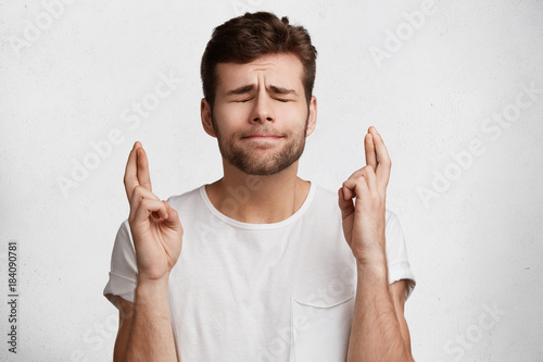 Unshaven brunet male closes eyes with big hope, crosses fingers as believes in something important, wears casual t shirt, isolated over white background. Hopeful handsome man dreams or expects