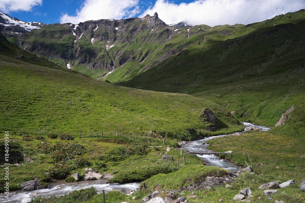 Landscape Picture on the stream or small river with rapids in the walley in the Alps mountains in austria in tirol region. Alpine meadow in summer, sunny day, blue sky, green grass and white clouds.