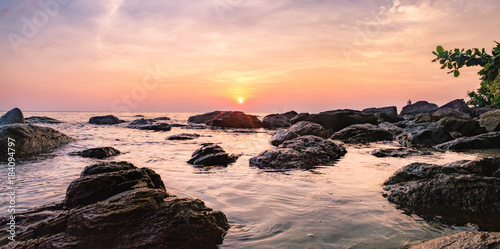 Tropical colourful sunset on the beach of Koh Chang island  Gulf of Siam  Thailand. Panorama view of dark night sea with protruding stones. A man sits on the rocks and admires the sunset