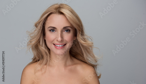 Beauty concept. Portrait of happy attractive middle-aged woman is standing with naked shoulders and looking at camera with wide smile. Isolated background with copy space in the right side photo