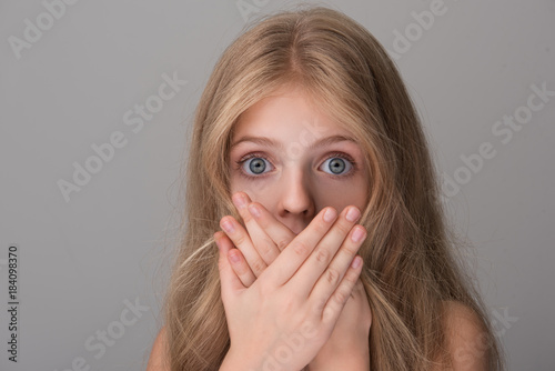 Close-up portrait of shocked little girl with long hair and naked shoulders  is looking at camera with wide-eyed while covering her mouth by hands.  Isolated background Stock Photo | Adobe Stock