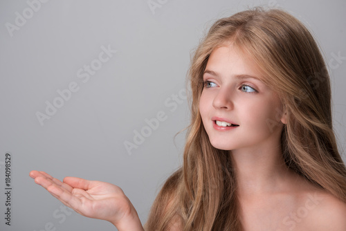Look here. Joyful pretty girl with long hair is looking aside with smile while holding out her hand palm up. Isolated background with copy space in the left side