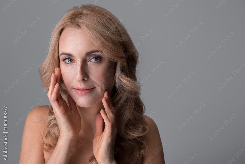 Natural attractiveness. Portrait of charming naked middle-aged woman is touching her face while standing and looking at camera temptingly. Isolated background. Copy space Natural attractiveness. Portrait of charming naked middle-aged woman is touching her face while standing and looking at camera temptingly. Isolated background. Copy space