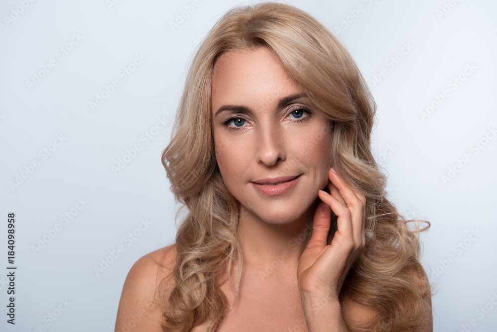 Perfect skin. Portrait of attractive elegant middle-aged naked woman is looking at camera with slight smile while touching her face with tenderness. Isolated background Perfect skin. Portrait of attractive elegant middle-aged naked woman is looking at camera with slight smile while touching her face with tenderness. Isolated background