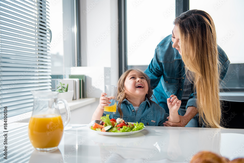 My little baby. Happy asian woman is expressing tenderness to her daughter while standing in kitchen. Girl is drinking juice and laughing