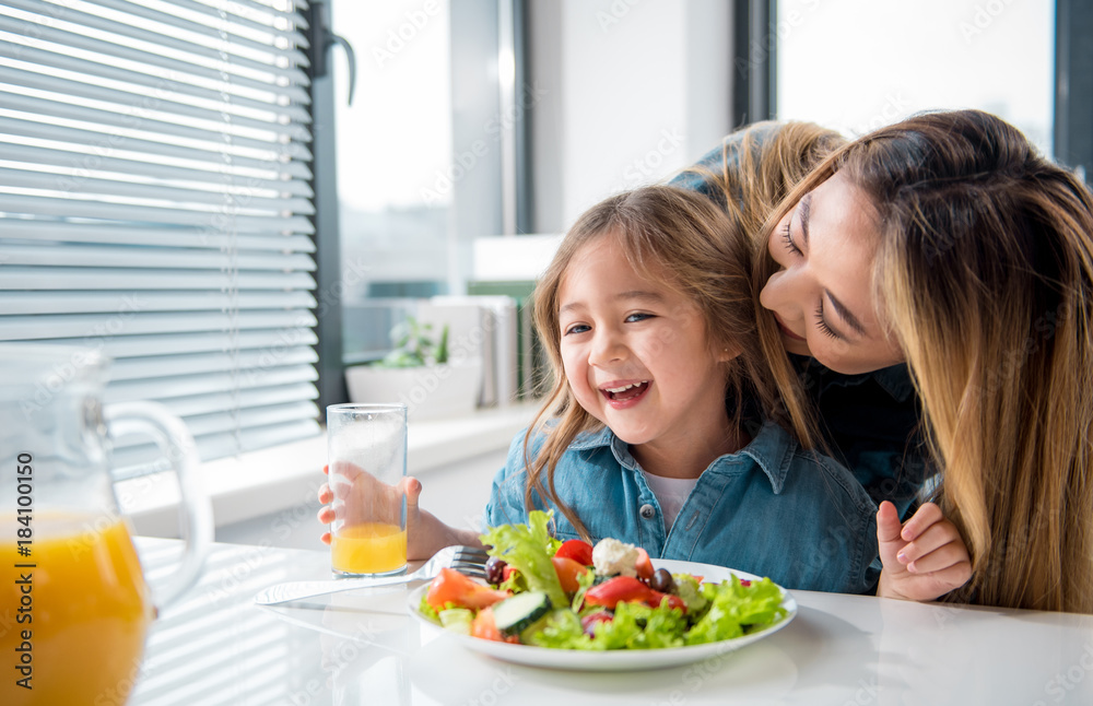 Cute asian girl is having healthy breakfast in kitchen. Her mother is kissing her with love. Child is sitting at table and smiling