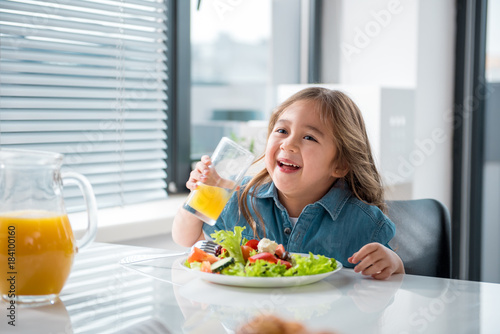 Portrait of pretty asian girl drinking healthy juice with pleasure. She is laughing while sitting at table near plate of salad