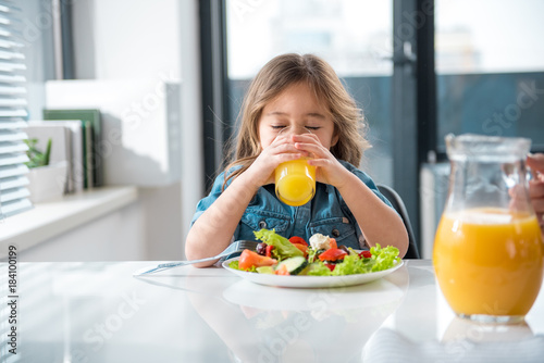 Thirsty little female child is drinking juice with enjoyment. She is sitting at table near plate of salad