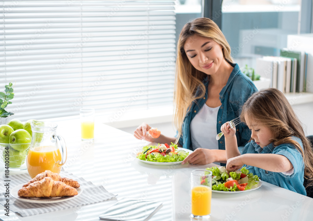 Side view of kind mother teaching her little daughter to lead healthy lifestyle. She is looking at girl taking piece of vegetable from plate and smiling. Copy space Side view of kind mother teaching her little daughter to lead healthy lifestyle. She is looking at girl taking piece of vegetable from plate and smiling. Copy space