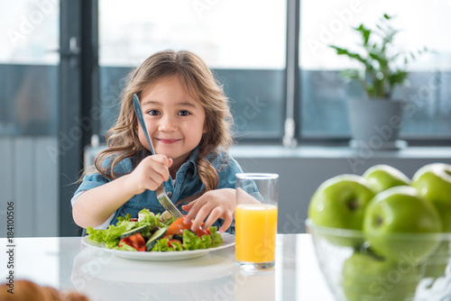 Portrait of pretty little female child having healthy breakfast. She is holding fork under the chopped vegetables and smiling photo