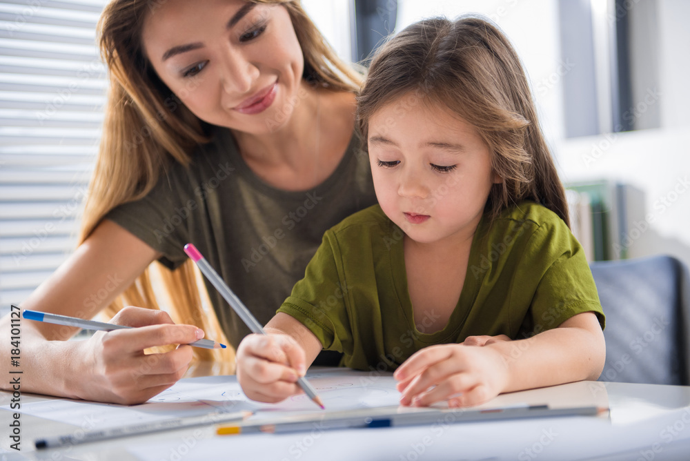 Portrait of joyful girl drawing picture with her mother with interest. She is holding pink pencil while sitting at table at home Portrait of joyful girl drawing picture with her mother with interest. She is holding pink pencil while sitting at table at home