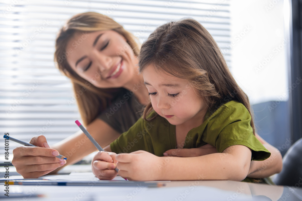 Drawing picture for loving mommy. Portrait of cute little child creating art on paper by pencil. Her mother is watching the process with excitement and smiling