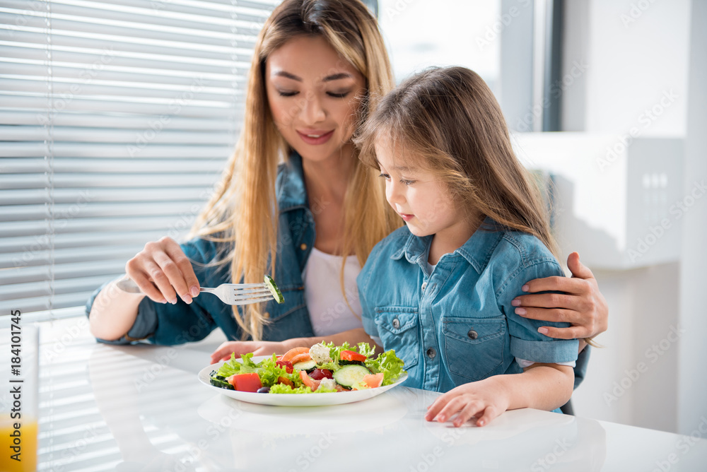 Pretty asian girl is eating fresh salad with help of her mother. She is looking at piece of cucumber on fork with interest