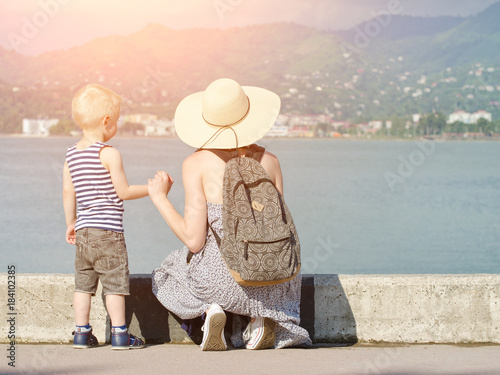 Mom and her son at the pier on the sea background, lighthouse and mountains in the distance. Back view