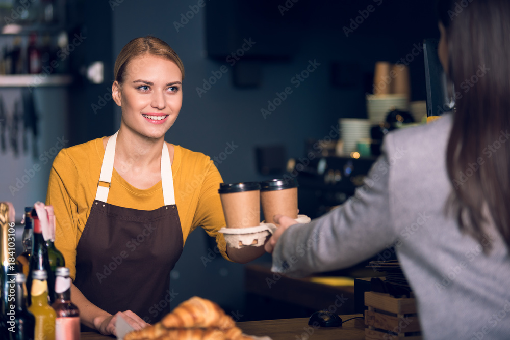Portrait of cheerful female barista giving two cups of delicious coffee to visitor. She standing at counter in cozy confectionary shop. Work concept