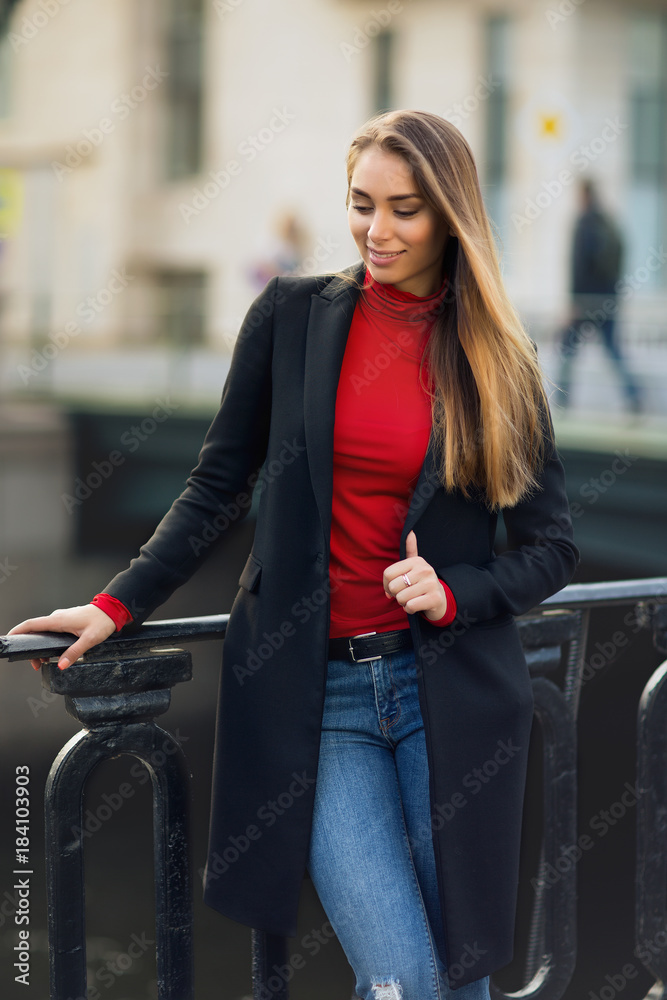 A portrait of young smiling woman with brown long hear, in black coat and  red top, blue jeans, standing by the fence on the embankment with cityscape  on background / Stock Photo